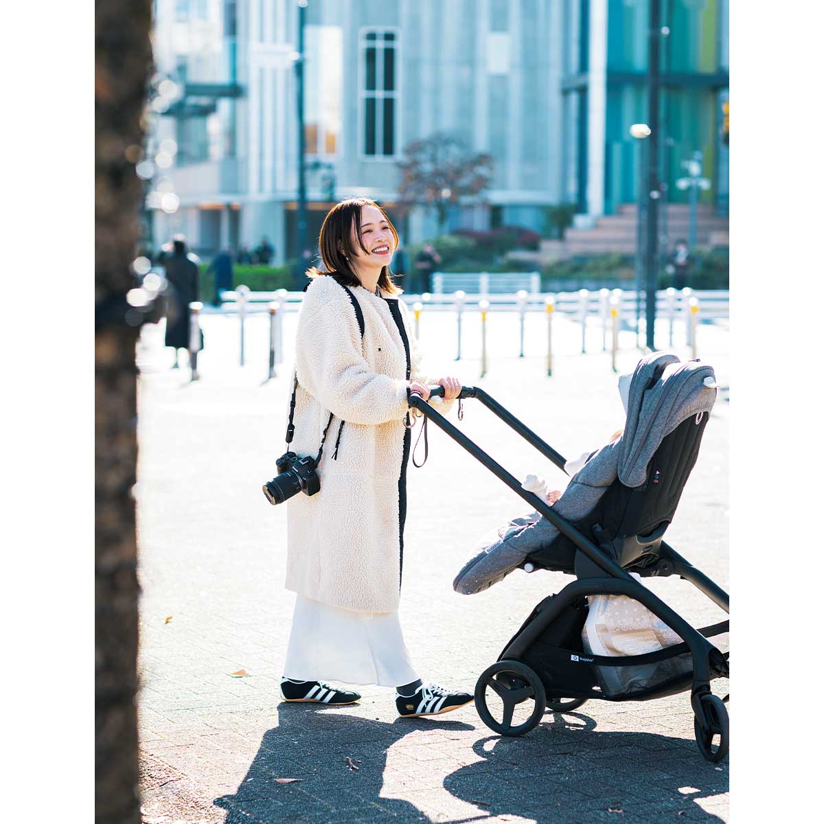 白のボアコートとスカートに黒スニーカーを履いて肩からカメラをさげてベビーカーを押すママ